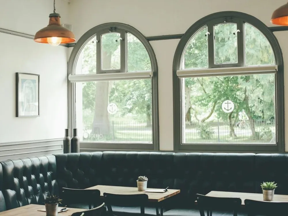 An empty café with tall arched windows, a dark green leather banquette and small tables