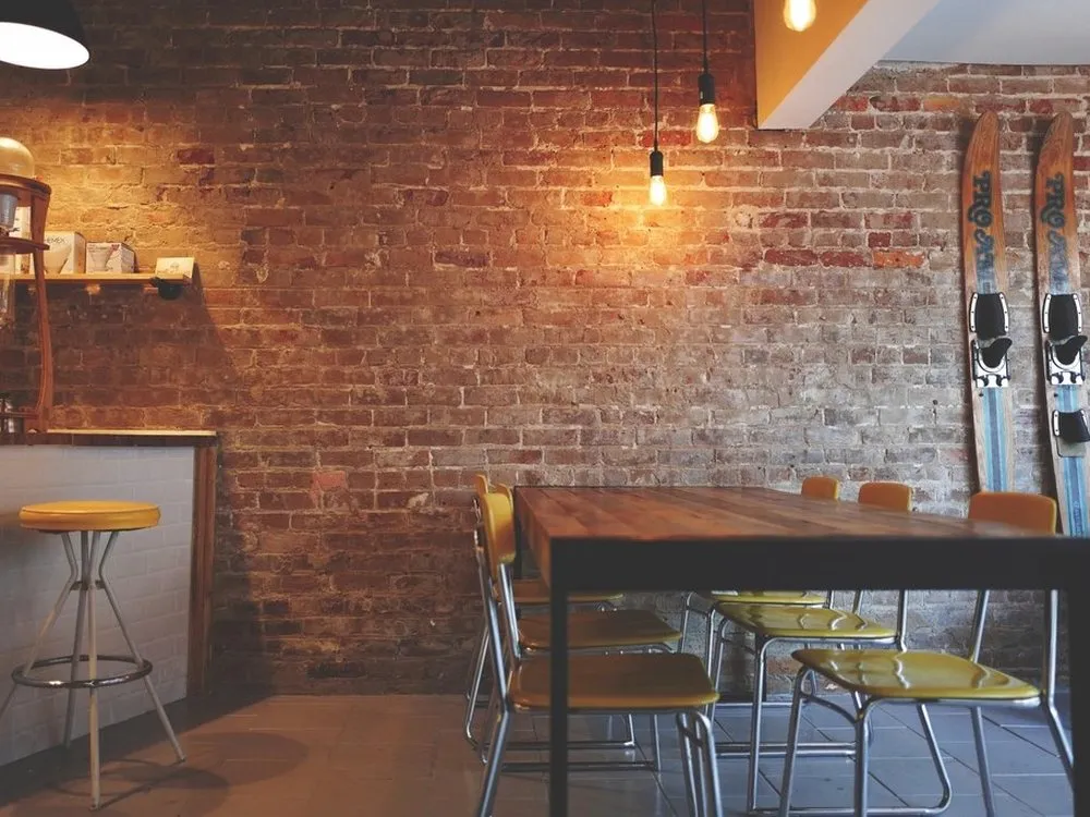 An empty café with an exposed brick wall, a long wooden table and bar stools under pendant lights