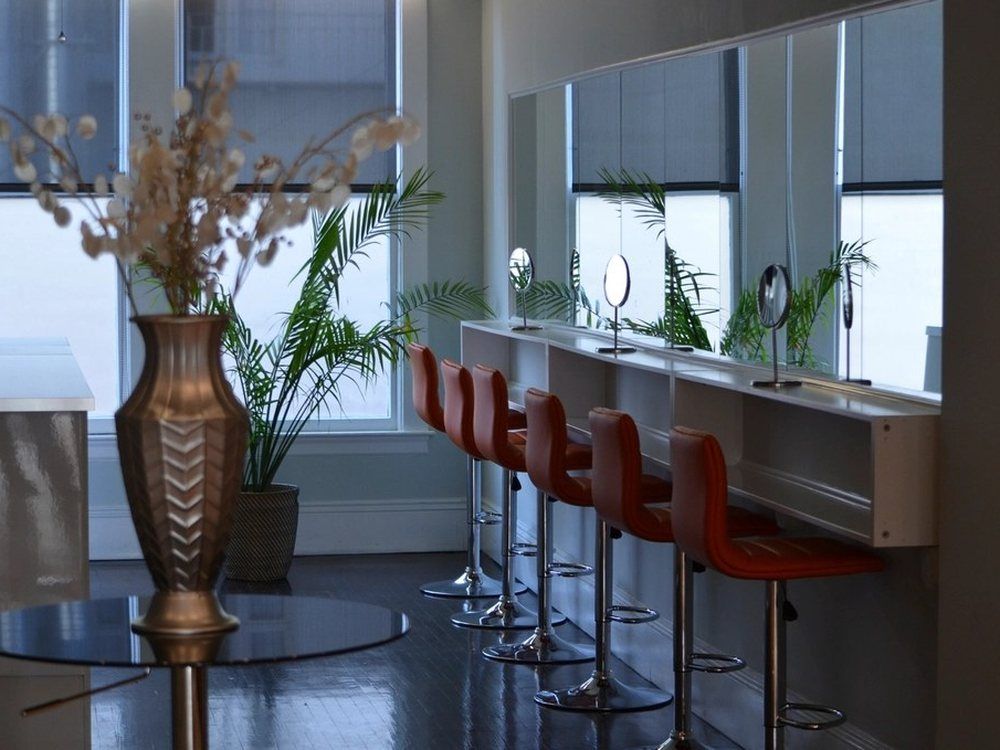 An empty salon with a row of red chairs facing mirrors along a window wall